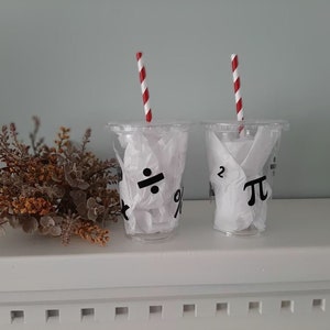 May include: Two clear plastic cups with red and white striped straws. The cups are filled with white tissue paper and have black mathematical symbols printed on them. The symbols are a division sign, a percentage sign, a superscript 2, and the Greek letter pi.