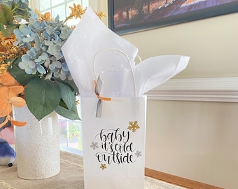 May include: White gift bag with a gold and black snowflake design and the text "Baby it's cold outside". The bag is filled with white tissue paper and tied with a black ribbon.