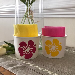 May include: Two white paper cups with a hibiscus flower design. One cup has a red flower and the other has a yellow flower. The cups are sitting on a table with a linen tablecloth.