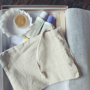 Może przedstawiać: Biała bawełniana torba ze sznurkiem, zawierająca mały pojemnik z złotym brokatem, dwie tubki farby i dwa drewniane patyczki. Przedmioty znajdują się w białym pudełku na brązowym drewnianym tle.