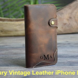 Könnte beinhalten: Eine braune iPhone-Hülle aus Leder mit weißen Nähten und einem Messingverschluss. Die Hülle hat einen Vintage-Look und zeigt die Initialen "GMA". Das Design ist klassisch und elegant.