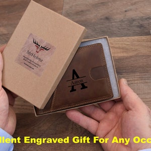 K&ouml;nnte beinhalten: Eine braune Ledergeldb&ouml;rse mit einer personalisierten gravierten Initial "A" und dem Namen "Aaron" auf der Vorderseite. Die Geldb&ouml;rse befindet sich in einer braunen Pappbox mit einem wei&szlig;en Etikett, auf dem "Mr.Holmz" steht.