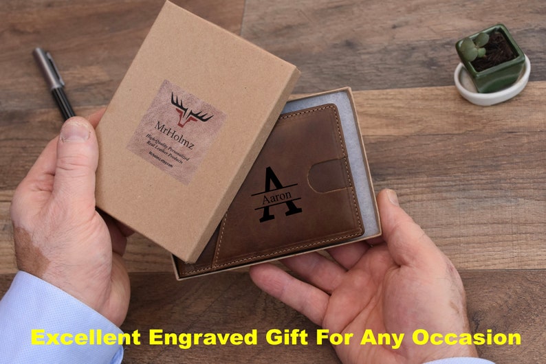 K&ouml;nnte beinhalten: Eine braune Ledergeldb&ouml;rse mit einer personalisierten gravierten Initial "A" und dem Namen "Aaron" auf der Vorderseite. Die Geldb&ouml;rse befindet sich in einer braunen Pappbox mit einem wei&szlig;en Etikett, auf dem "Mr.Holmz" steht.