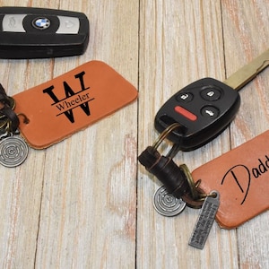 Könnte beinhalten: Zwei Leder-Schlüsselanhänger mit Metallringen und Anhängern. Ein Schlüsselanhänger hat die Initialen "T.W." mit dem Namen "Wheeler" darunter. Der andere Schlüsselanhänger hat das Wort "Daddy" in Kursivschrift geschrieben.