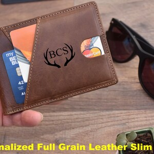 K&ouml;nnte beinhalten: Eine schlanke Geldb&ouml;rse aus braunem Leder mit einem personalisierten Monogramm "BCS" mit Geweih. Die Geldb&ouml;rse hat ein Kartenfach und einen Geldclip.