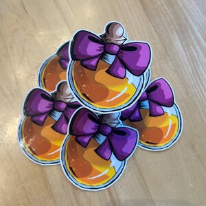 May include: A stack of stickers featuring a cartoon potion bottle design. The bottles are round with a light brown stopper and filled with orange liquid. Each bottle has a large purple bow. The stickers are on a light brown surface.