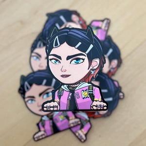 May include: A sticker featuring a cartoon illustration of a girl with black hair, pink jacket, and black cat ears. She is wearing a black choker with the word "HELLS" on it. The sticker has a pink and yellow color scheme.