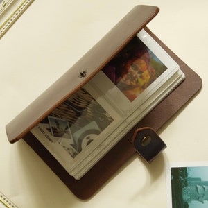 May include: Brown leather photo album with a flap closure and a strap. The album is open to reveal white pages with clear plastic sleeves for holding photos. The text "Instax Square" is printed on the front cover.