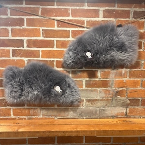 May include: Two gray, fluffy sheepskin slippers are displayed against a red brick wall. Each slipper has a small tag attached. The slippers are positioned as if they are hanging on the wall, showcasing their shape and texture.