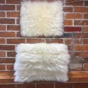 May include: Two fluffy, square, off-white sheepskin cushions. They are set against a rustic red brick wall and wooden shelf. The sheepskin has a soft, shaggy texture.