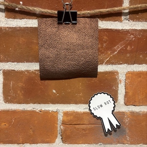 May include: A bronze-coloured leather swatch, clipped to a rope with a black binder clip, hangs against a brick wall. A white ribbon-shaped sticker with the words "SLOW ROT" in black lettering is also attached to the wall.