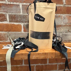 May include: A kraft paper bag with a clear window and the text "SLOW ROT" on a black tag. The bag contains black items and is surrounded by black, white, and gray leather strips. The background is a brick wall.