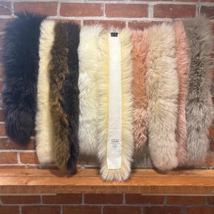 May include: A collection of long-haired faux fur strips in various colours including black, dark brown, cream, white, light pink, and grey. A central white fabric tag with the text SLOW SUIT is visible on one of the cream-coloured fur strips.