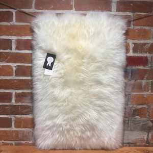May include: Cream-coloured rectangular sheepskin rug with long, fluffy fibres. The rug is displayed against a rustic brick wall and a wooden shelf. A small tag is attached to the rug.