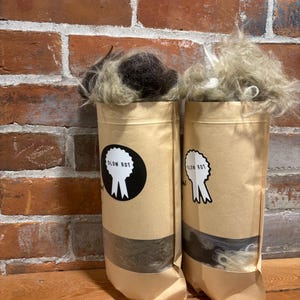 May include: Two cylindrical paper bags filled with fluffy, light-coloured fibre. Each bag has a black and white circular sticker with the text "SLOW ROT" and a tree-like graphic. The bags are on a wooden surface against a brick wall.