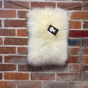 May include: A rectangular off-white faux fur rug with long, dense fibers. A black rectangular tag is attached to the upper right corner, featuring a white ribbon icon and the text "100% GENUINE SHEEPSKIN" printed in white capital letters.
