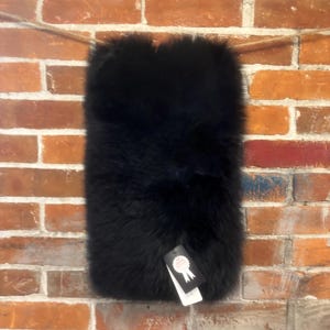 May include: A rectangular, black fur item hangs against a brick wall. The fur appears soft and plush. A white tag with black text is attached to the bottom of the fur item. The brick wall has a mix of red, brown, and white bricks.