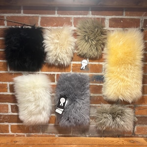 May include: A collection of seven rectangular faux fur swatches in various colours including black, cream, beige, yellow, white, and grey. Each piece features long, shaggy synthetic fibres. One grey swatch has a black tag with a white ribbon icon.