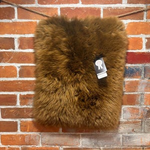 May include: A rectangular, brown sheepskin rug with a thick, shaggy texture. The rug is hanging against a brick wall with a wooden beam at the bottom. A tag is attached to the rug.