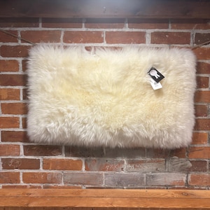 May include: A rectangular, off-white sheepskin rug with a thick, fluffy texture. The rug is displayed against a red brick wall, with a wooden shelf below. A small tag is attached to the rug.