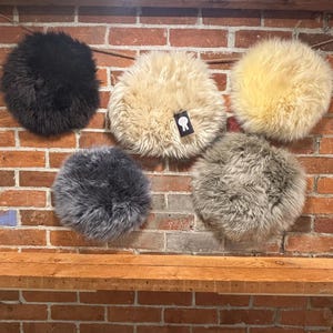 May include: Five circular sheepskin rugs in black, cream, light gray, dark gray, and beige. Each rug features long, dense, fluffy fur. One cream-colored rug includes a small black tag with a white ribbon icon attached to the edge of the fur.