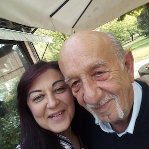 Puede incluir: Una foto de cerca de un hombre y una mujer sonriendo a la c&aacute;mara. El hombre tiene un bigote blanco y lleva un su&eacute;ter azul oscuro. La mujer tiene el pelo largo y oscuro y lleva una camisa con estampado blanco y negro.
