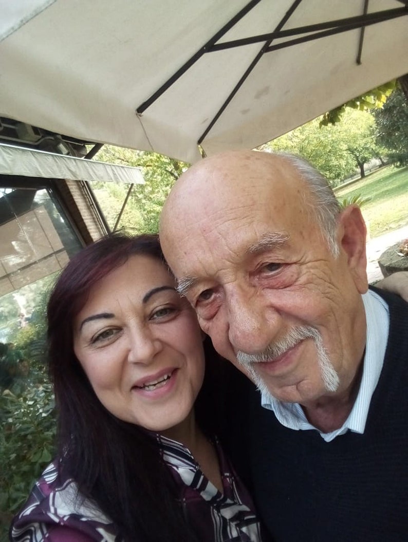 Puede incluir: Una foto de cerca de un hombre y una mujer sonriendo a la c&aacute;mara. El hombre tiene un bigote blanco y lleva un su&eacute;ter azul oscuro. La mujer tiene el pelo largo y oscuro y lleva una camisa con estampado blanco y negro.