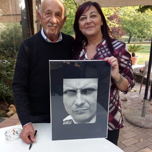 Puede incluir: Dos personas est&aacute;n de pie frente a una mesa blanca. La persona de la izquierda sostiene una pluma negra. La persona de la derecha sostiene una foto en blanco y negro enmarcada de la cara de un hombre. La foto est&aacute; en un marco negro con un passe-partout blanco.