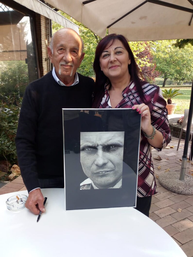 Puede incluir: Dos personas est&aacute;n de pie frente a una mesa blanca. La persona de la izquierda sostiene una pluma negra. La persona de la derecha sostiene una foto en blanco y negro enmarcada de la cara de un hombre. La foto est&aacute; en un marco negro con un passe-partout blanco.