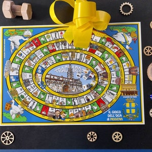 Op de afbeelding: Een kleurrijk spelbord met een geel lint erop. Het bord heeft een cirkelvormig parcours met nummers en afbeeldingen van ganzen. Het spel heet "Il Gioco Dell'Oca di Modena".