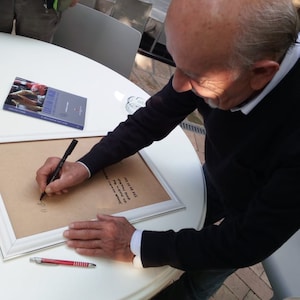 Puede incluir: Una persona firmando un trozo de papel enmarcado blanco con un bol&iacute;grafo negro. El papel tiene escritura negra. La persona lleva una chaqueta negra y pantalones vaqueros azules. Hay un bol&iacute;grafo rojo y un libro sobre la mesa.