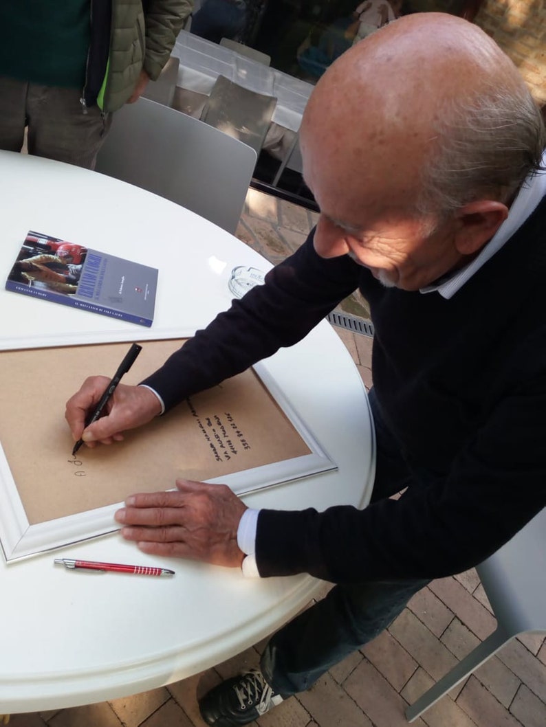 Puede incluir: Una persona firmando un trozo de papel enmarcado blanco con un bol&iacute;grafo negro. El papel tiene escritura negra. La persona lleva una chaqueta negra y pantalones vaqueros azules. Hay un bol&iacute;grafo rojo y un libro sobre la mesa.