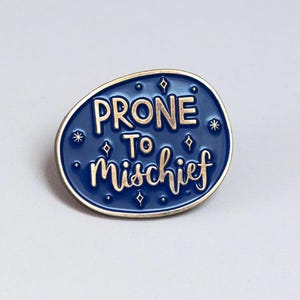 Peut inclure: Épingle en émail bleu avec l'inscription dorée « PRONE TO Mischief ». L'épingle a une bordure dorée et est ornée de petites étoiles et de diamants. Accessoire idéal pour vestes, sacs ou chapeaux.