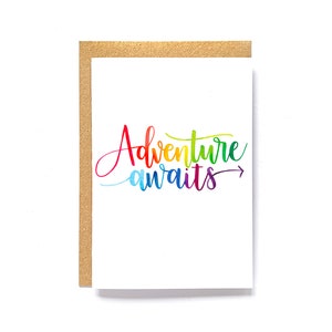 Peut inclure: Carte de vœux blanche avec l'inscription "Adventure awaits" en écriture multicolore. Le texte, allant du rouge au violet, est orné d'une petite flèche. La carte est encadrée d'un cadre doré texturé.