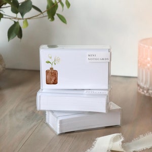 May include: A stack of white mini notecards in a clear plastic box with a watercolor illustration of a brown glass vase with white flowers. The box has a white label that says "MINI NOTECARDS".