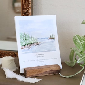 May include: A white card with a watercolor landscape scene of a shoreline with trees and water, held in a small wooden block. The card has the word "sovereign" and text from Psalm 103:19.