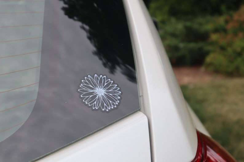 May include: A white outline of a daisy flower sticker on a black car window.