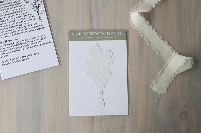 May include: White and clear car window decal featuring a floral design. The decal is packaged in a clear plastic sleeve with a white card that reads "CAR WINDOW DECAL" and "WHITE & CLEAR".