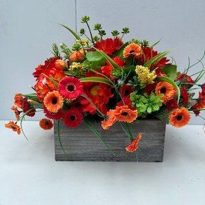 May include: A colourful floral display in a wooden box. The arrangement includes red and orange gerbera daisies, green leaves, and small yellow flowers. The box is made of grey, weathered wood.