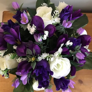 Può includere: Una composizione floreale con rose artificiali viola scuro e bianche, fiori più piccoli color viola chiaro e minuscoli fiori bianchi di riempimento. Il bouquet include foglie e steli verde scuro, creando un display denso e arrotondato.