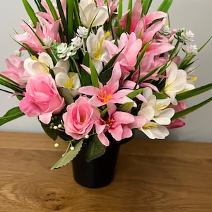 Pode incluir: Um arranjo floral vibrante em um vaso preto. O buquê apresenta lírios rosa, rosas e flores brancas com centros amarelos, complementados por folhas verdes e grama. O arranjo está em uma superfície de madeira.