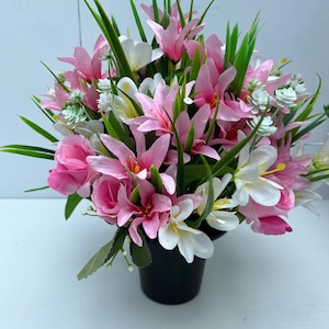 May include: A vibrant floral arrangement in a black pot. The arrangement features pink lilies, white flowers, and green foliage. The flowers are clustered together, creating a full and lush appearance. The pot is round and sits on a white surface.