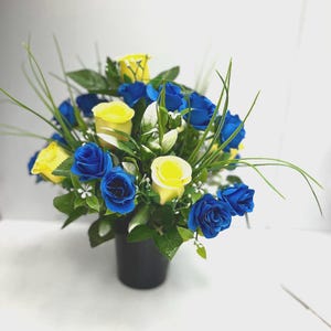 May include: A floral arrangement featuring vibrant blue and yellow roses nestled in a black cylindrical container. The bouquet includes green leaves and thin, blade-like grass accents interspersed among the colorful flower heads.