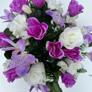 Peut inclure: Un bouquet de fleurs artificielles comprenant des tulipes violettes, des roses blanches et des orchidées lavande. L'arrangement comprend de petites fleurs de remplissage blanches et des feuilles vertes, créant un affichage vibrant et coloré.