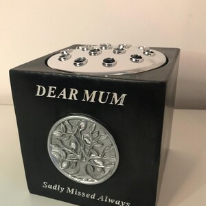 Pode incluir: Vaso memorial quadrado preto com um design floral prateado e as palavras "DEAR MUM" e "Sadly Missed Always". A parte superior tem uma placa prateada com vários orifícios para caules de flores.