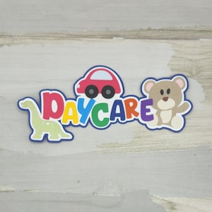 May include: A colorful daycare-themed cutout with the word "DAYCARE" in rainbow colors. The design includes a red car, a light green dinosaur, and a brown teddy bear. The cutout has a blue border and is set against a light wood-grain background.