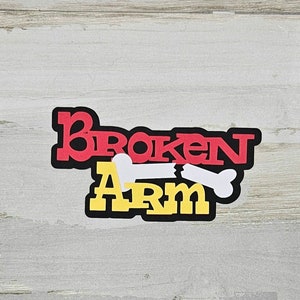 May include: A sticker with the words "BROKEN ARM" in red and yellow block letters, outlined in black. A white bone graphic is broken in the middle of the text, suggesting an injury. The sticker is on a light gray and white wood-grain background.