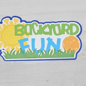 May include: A colorful die-cut sign with the words "BACKYARD FUN" in green and blue, featuring a yellow sun, an orange ball, and green grass. The sign has a blue border and is on a light gray surface.