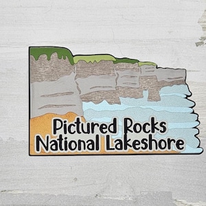 May include: A colorful enamel pin featuring a stylized illustration of a cliffside with water crashing against the rocks. The text "Pictured Rocks National Lakeshore" is printed in black on a yellow background.