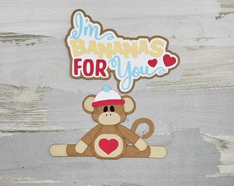 May include: A brown monkey with a blue and white hat is sitting with its legs spread out. The monkey has a red heart on its chest. Above the monkey is a brown sign with the text "I'm Bananas For You" in yellow, blue, and red letters. There are two red hearts on the sign.